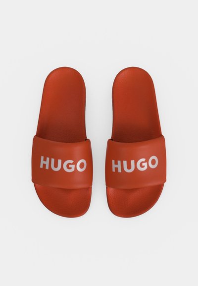 Двойка червени сандали с широки презрамки с бял надпис "HUGO" на всяка презрамка на едноцветен фон.