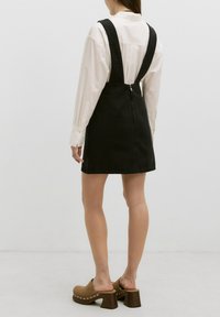 Zwarte spijkeroverall jurk met verstelbare banden, getailleerd ontwerp en een zijrits. Gecombineerd met bruine platformklompen met metalen accenten.