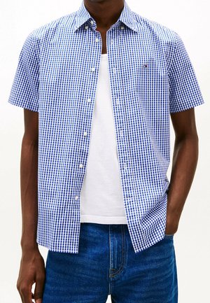 Mann trägt ein kurzärmliges blau-weiß kariertes Hemd über einem weißen T-Shirt und blauen Jeans, steht mit einer Hand in der Tasche.