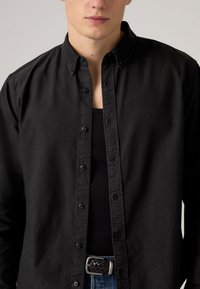 Μαύρο πουκάμισο με κουμπιά από βαμβάκι, διαθέτει κλασικό γιακά, μακριά μανίκια και διακριτικό λογότυπο. Φορεμένο πάνω από μαύρο τοπ χωρίς μανίκια.