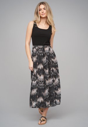 Femme aux longs cheveux blonds portant un haut noir sans manches et une jupe longue fluide avec un motif noir et gris, ainsi que des sandales, debout sur un fond gris.