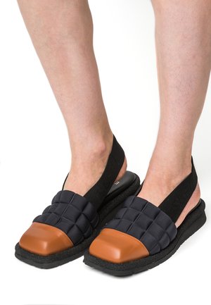 Piedi che indossano sandali trapuntati neri con puntali in pelle marrone e cinturini elastici neri sul tallone su sfondo bianco.