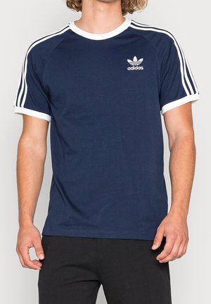 Homme portant un t-shirt Adidas bleu marine avec des bandes blanches sur les manches et un logo sur la poitrine, assorti à un pantalon noir.
