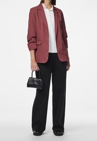 Blazer color ruggine con spalle strutturate, abbinato a una camicia bianca e pantaloni neri a gamba larga. Porta una piccola borsa nera in pelle con motivo coccodrillo.