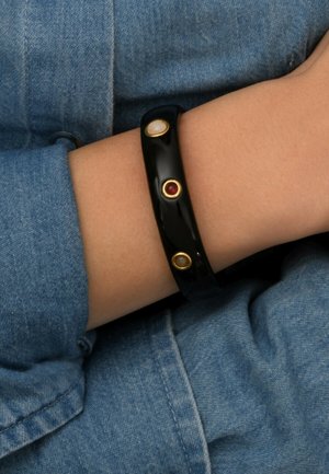 Bracelet noir brillant avec trois pierres décoratives : une rouge et deux blanche cassé, chacune sertie d'accents en ton or. Porté au poignet sur un tissu bleu.