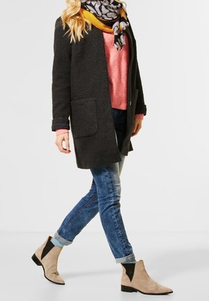 Manteau noir texturé avec deux poches avant, superposé à un pull rose et un jean bleu. Bottines beiges avec des accents noirs complètent le look.