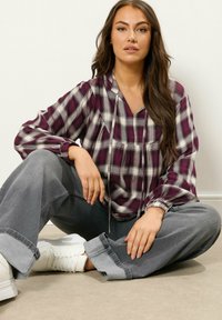 Pruimkleurige en witte geruite blouse met een v-hals, trekkoorddetails en pofmouwen; gecombineerd met losse grijze jeans en witte sneakers.