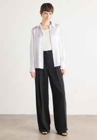 Hvit skjorte med knapper laget av glatt stoff, brukt over en tettsittende beige tank top, kombinert med høyt midjebånd, svarte vide bukser.