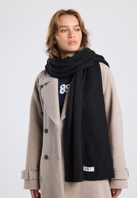 Zwarte sjaal van zachte stof, gedrapeerd om de nek, met een gladde textuur en een klein logo-label zichtbaar aan één uiteinde. Draagt een lichtgrijze jas.