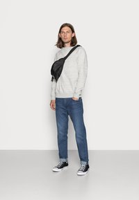 Riñonera negra llevada de forma cruzada, sudadera gris con puños acanalados, jeans azul ajustados y zapatillas negras con suelas blancas.