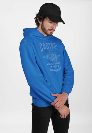 Blå hættejakke med en frontlomme og ribbede manchetter, prydet med et gråt grafisk logo. Modellen bærer en sort kasket og sorte bukser.