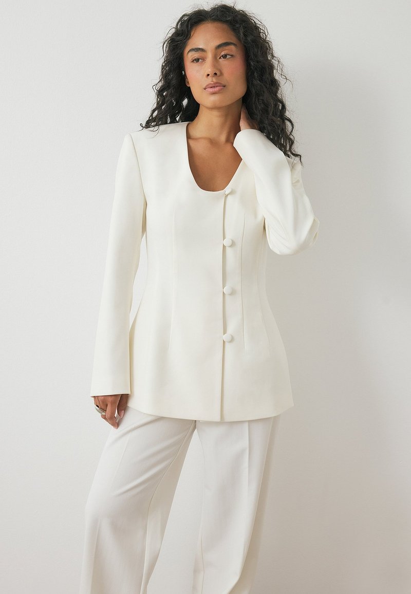 Valkoinen muokattu bleiseri, jossa on neliömäinen kaulus, pitkät hihat ja kolmen napin suljenta. Liukas kangas, jossa on jämäkät hartiat ja istuva siluetti.