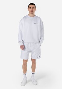 Sudadera blanca de algodón con cuello redondo y un logo negro, combinada con pantalones cortos blancos de algodón que presentan un logo negro en el dobladillo. Zapatillas blancas y calcetines.