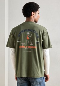 T-shirt de algodão verde com um design impresso nas costas com "CLUBE DE CORRIDA" e uma personagem vestindo uma blusa vermelha. Corte casual, mangas curtas.