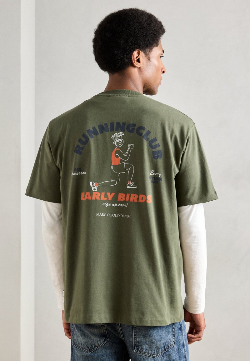 T-shirt de algodão verde com um design impresso nas costas com "CLUBE DE CORRIDA" e uma personagem vestindo uma blusa vermelha. Corte casual, mangas curtas.