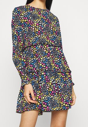 Vrouw die een zwarte jurk met lange mouwen draagt met een meerkleurig klein bloemenpatroon en gelaagde ruches bij de taille en zoom.