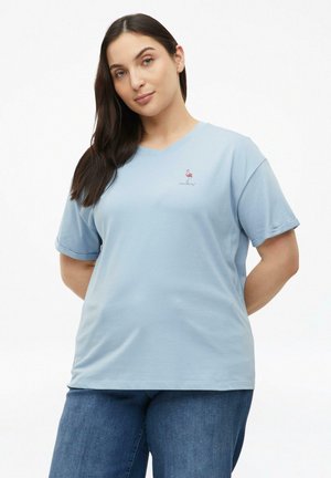 Frau mit langen dunklen Haaren, die ein hellblaues T-Shirt mit einem kleinen rosa Flamingo-Motiv und blaue Jeans trägt, steht mit den Händen hinter dem Rücken.