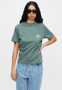 Roheline puuvillane T-särk ümmarguse kaelusega ja lühikeste varrukatega, millel on vasakul rinnal tasku ja väike brändi logo kant. Kandmisel helesinised triibulised teksad.