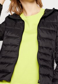 Černá péřová bunda s horizontálními prošívanými vzory, vybavená zipem a připojenou kapucí, nošená přes jasně neonově zelené tričko.