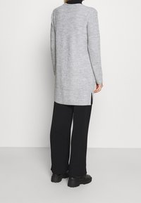 Osoba ubrana w długi, szary sweter w prążki, czarne spodnie i czarne masywne buty, stoi tyłem na jednolitym jasnym tle.