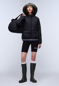 Black quilted jacket with pockets, green wide-brimmed hat, black shorts, and green knee-high boots. Model holding a black duffel bag.