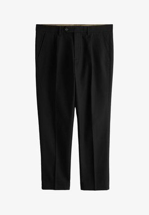 Man draagt zwarte geplooide broek en een wit overhemd met lange mouwen en knopen, met linkerhand rustend naast de broek.