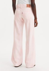 Personne portant un jean Levi's léger rose clair à taille haute et jambes larges, avec des poches arrière et une étiquette en cuir de la marque, se tenant devant un fond blanc.