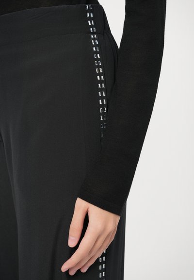 Czarne spodnie o gładkiej fakturze mają pionowy rząd srebrnych kwadratowych ćwieków wzdłuż boku, co podkreśla ich ogólny design.