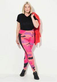 Flerfärgade leggings med rosa, lila och svarta mönster, ihop med en svart V-ringad skjorta och en röd jacka, fullbordat med svarta sneakers.