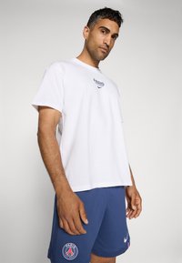 Hvid bomulds T-shirt med "PARIS" logo på brystet, parret med blå shorts med et rundt PSG logo og et Nike swoosh emblem.