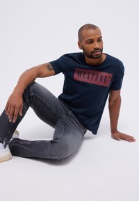 Tamsiai mėlynas medvilninis marškinėliai su dideliu bordo spalvos stačiakampio formos logotipu, ant kurio užrašyta "MUSTANG." Suderinti su pilkiem siaurais džinsais ir baltais sportbačiais.