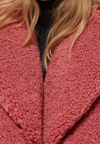 Zewnętrzna odzież z polaru w delikatnym odcieniu różu, z teksturowanym wykończeniem i szerokimi klapami, założona na czarny top. Zdjęcie z bliska podkreśla detale tkaniny.