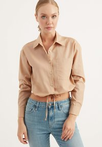 Camicetta beige corta con colletto, bottoni frontali e taschino sul lato sinistro del petto. Tessuto morbido e liscio con dettaglio a fiocco all'orlo.