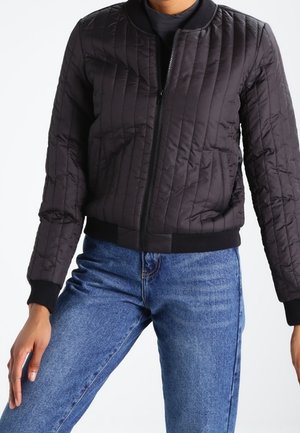 Frau trägt eine schwarze gesteppte Bomberjacke mit gerippten Bündchen und Saum, kombiniert mit blauen, hoch taillierten Jeans.