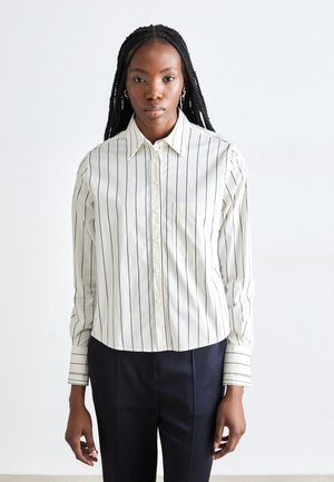 Camicia a righe bianche con colletto, caratterizzata da sottili righe verticali nere e una tasca sul petto. Il materiale sembra leggero e liscio.