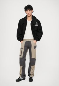 Black jacket with a front pocket, white t-shirt, beige and gray patchwork pants with pockets, and black athletic shoes with white stripes.