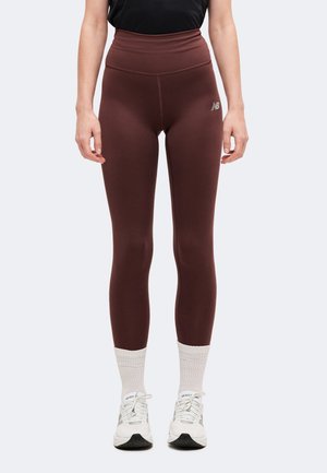 Femme portant des leggings marron foncé taille haute, des chaussettes blanches et des chaussures de sport blanches, debout devant un fond clair uni.