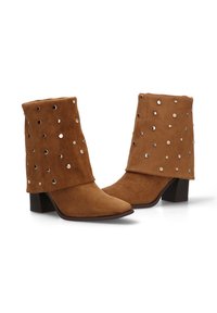 Botas de tobillo de ante marrón con un puño doblado que presenta tachuelas metálicas circulares, punta afilada y un tacón bajo y cuadrado.