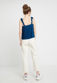 Námořnická modrá, žebrovaná tílko s ramínky ve stylu mašle, doplněné o krémové, mírně opotřebované džínové kalhoty rovného střihu a bílé tenisky.