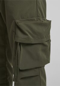 Primer plano del bolsillo de pantalones cargo verde oliva con solapa y detalle de cordón en el lateral de la pierna.