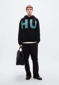 Μαύρο φούτερ με μεγάλο γαλάζιο γραφικό «HU», συνδυασμένο με μαύρο παντελόνι φόρμας με μικρό λογότυπο. Το μοντέλο κρατάει μια μαύρη τσάντα.