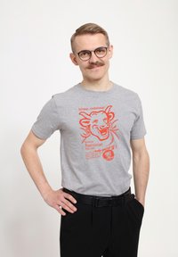Grå kortärmad T-shirt med orange grafisk design av en leende ko, med text och cirkulär logotyp. Bärs med svarta byxor och bälte.