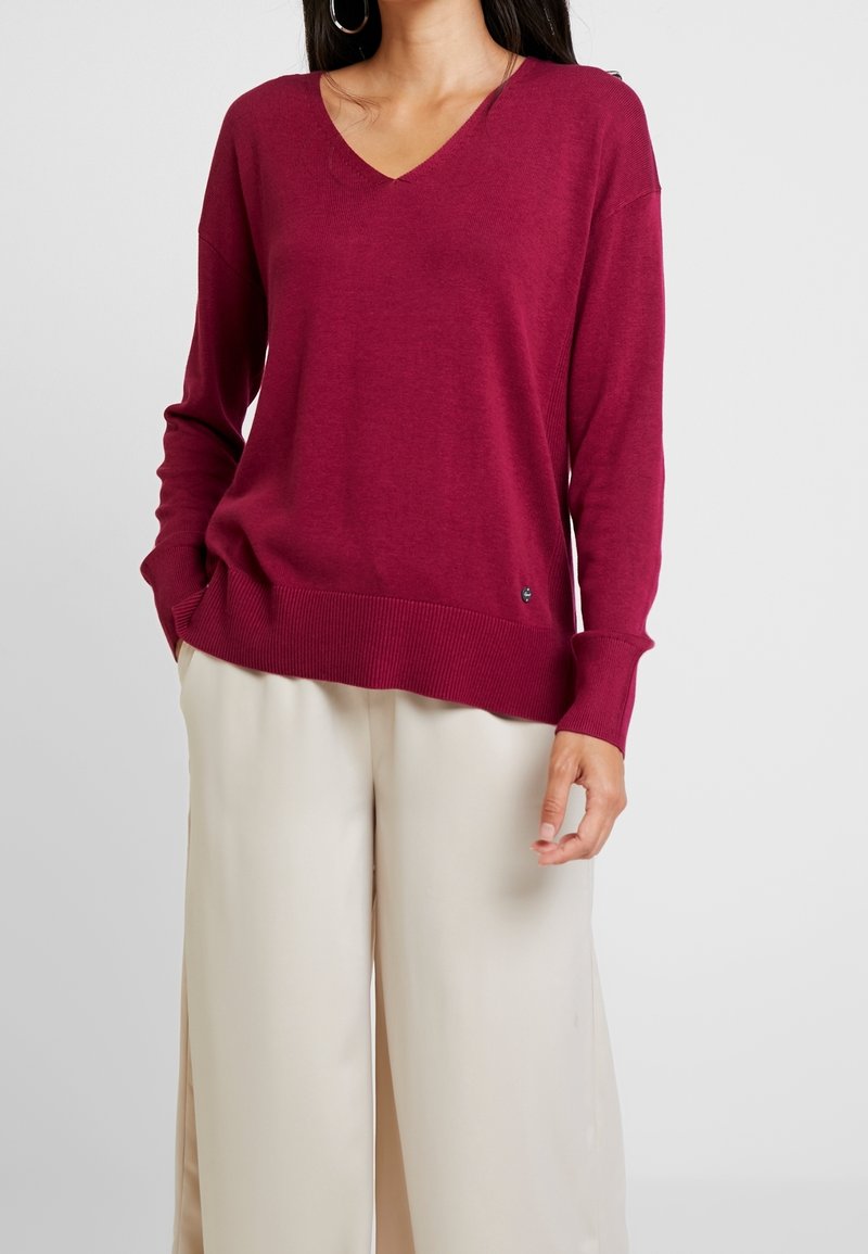 V-ringad tröja med långa ärmar i djup vinröd färg, tillverkad av mjukt, lätt tyg med ribbad fåll; bärs med cremefärgade byxor med vida ben.