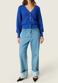 Blå stickad cardigan med V-ringning, knappstängning och texturerat mönster; ihop med höga ljusblå jeans och svarta spetsiga skor.