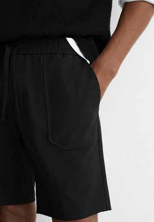 Person iført sorte shorts med elastisk talje og frontlomme, hånd i lommen, samt en sort sweater med hvid skjorte under.