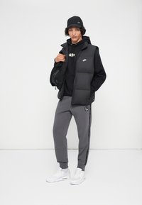 Jovem vestindo um boné preto da Nike, moletom preto, colete acolchoado preto, calças de moletom cinzas da Nike e tênis brancos, segurando uma bolsa preta sobre o ombro.