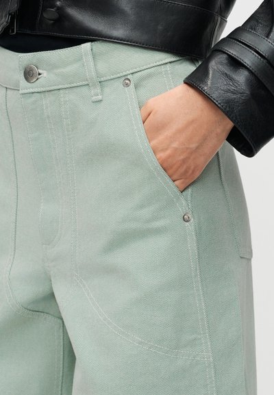 Χέρι στην τσέπη ανοιχτό πράσινου παντελονιού από τζιν, σε συνδυασμό με μανίκι μαύρου δερμάτινου μπουφάν εμφανές σε κοντινή λήψη.