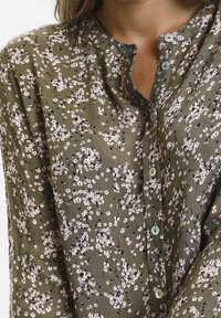 Olivgrünes Blusenshirt mit kleinen, verstreuten Blumenmustern in Rosa und Weiß. Verfügt über einen runden Halsauschnitt und eine Knopfverschluss. Faltenstruktur.