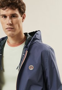 Marineblaue wasserdichte Jacke mit Kapuze, Frontreißverschluss und einem runden Logo-Patch. Über einem hellgrünen T-Shirt mit rundem Ausschnitt getragen.