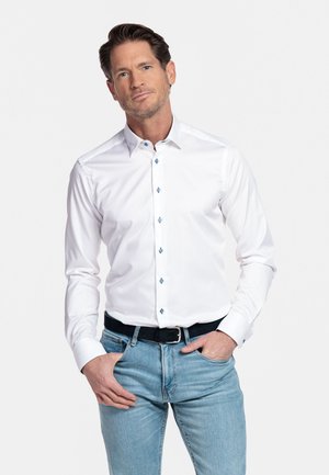 Mann mit kurzem braunem Haar, der ein weißes Hemd mit Knöpfen und hellblaue Jeans trägt, posiert mit den Händen in den Taschen.